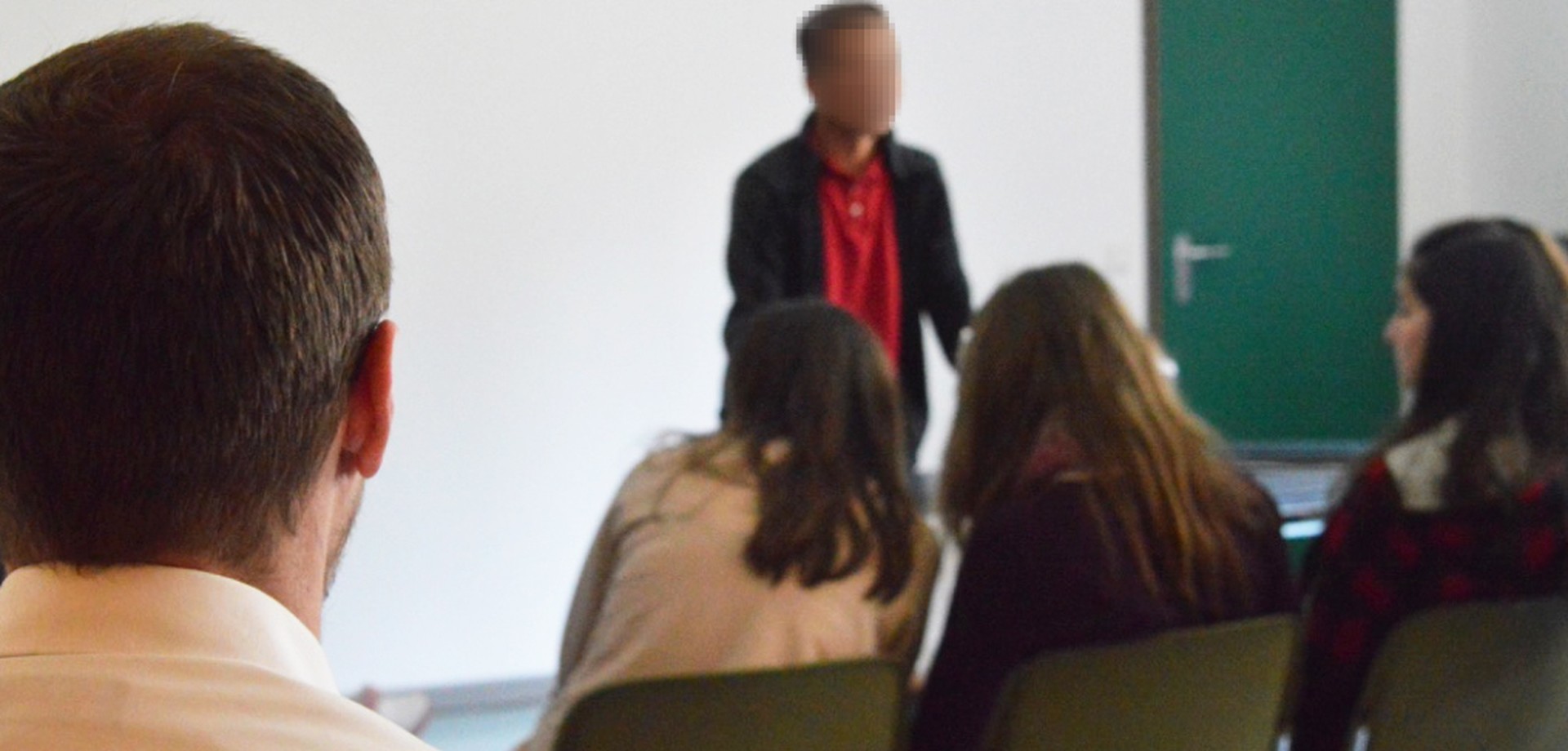 Ein Patient mit gepixelten Gesicht referiert vor Schülern, die in Stuhlreihen sitzen.