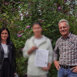 Zwei Männer und eine Frau schauen in die Kamera vor einer grünen Hecke.