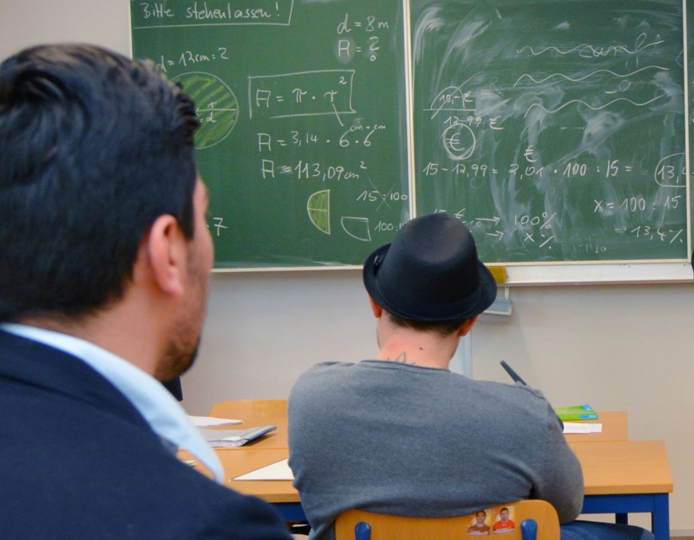 Zwei junge Männer sitzen auf Stühlen an Tischen vor einer mit mathematischen Formeln beschriebenen Tafel.