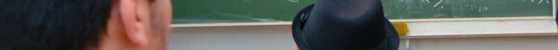 Zwei Männer sitzen, auf eine vollgeschriebene Tafel schauend, in einem Klassenraum.