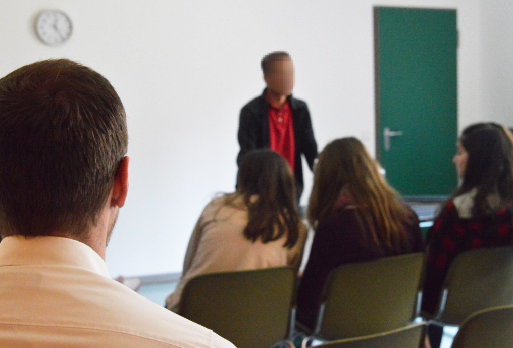 Ein Patient, der unkenntlich gemacht wurde, steht vor einer Gruppe sitzender Schülerinnen und hält einen Vortrag.